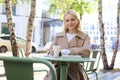 Portrait of stylish modern woman, sitting in an outdoor cafe, smiling and drinking coffee from takeaway cup, wearing Royalty Free Stock Photo