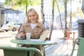 Portrait of stylish modern woman, sitting in an outdoor cafe, smiling and drinking coffee from takeaway cup, wearing Royalty Free Stock Photo