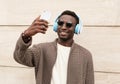 Portrait stylish smiling african man taking selfie picture by smartphone in wireless headphones listening to music wearing brown Royalty Free Stock Photo