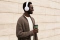 Portrait stylish smiling african man in wireless headphones listening to music with coffee cup wearing brown knitted cardigan Royalty Free Stock Photo