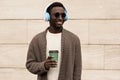 Portrait stylish smiling african man in wireless headphones listening to music with coffee cup wearing brown knitted cardigan