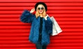 Portrait stylish smiling woman with shopping bags wearing blue faux fur coat, black round hat and sunglasses posing over red wall