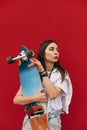 Portrait of stylish woman in casual clothes standing on red background with longboard in hands, looking aside with serious face