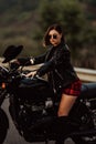 Portrait of stylish woman sitting on classic black retro-styled motorcycle while standing on road. Young lady in leather