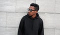 Portrait stylish young african man posing looking away wearing a black hoodie, sunglasses on a city street over gray wall Royalty Free Stock Photo