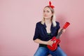 Portrait of stylish young blonde with ukulele. A woman with a headband on a pink background