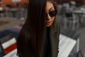 Portrait of a stylish and young brunette girl in an autumn green oversized coat and sunglasses sitting in an outdoor cafe.