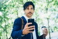 Portrait of stylish young businessman using smartphone for listining music while walking in city park and holding take Royalty Free Stock Photo