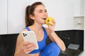 Portrait of stylish, young fitness woman, eating an apple and using mobile phone, holding smartphone, wearing sportswear Royalty Free Stock Photo