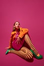 Portrait of stylish young girl in orange stripped tights, jacket and heel shoes, posing against magenta color studio