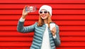 Portrait stylish young woman taking selfie by smartphone wearing a white hat, jacket on red background Royalty Free Stock Photo