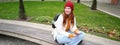 Portrait of stylish young woman, 25 years, sits on bench in park and uses mobile phone, reads online news, messages or Royalty Free Stock Photo