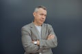 Portrait of successful aged gray haired businessman standing with crossed arms in stylish suit and luxury watch on gray