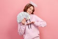 Portrait of successful curly haired teenage girl in hoodie with halo on head showing bunch of dollar banknotes and sincerely