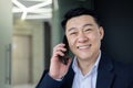 Portrait of successful mature asian businessman inside office at workplace, senior boss talking on phone, smiling and Royalty Free Stock Photo