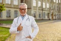 Portrait of successful senior male doctor in lab coat and glasses smiling at camera, showing thumbs up, standing Royalty Free Stock Photo