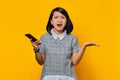Portrait of surprised asian woman holding mobile phone with confused and unhappy expression on yellow background Royalty Free Stock Photo