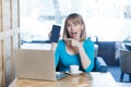 Portrait of surprised beautiful young blonde woman in blue t-shirt, sitting with laptop, holding and pointing mobile display and Royalty Free Stock Photo