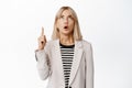 Portrait of surprised blond woman in suit pointing, looking up with fascinated, impressed face expression, checking out