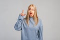 Portrait of surprised blond young woman in blue sweatshirt pointing finger and looking up with fascinated, impressed face