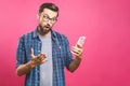 Portrait of a surprised casual man looking at mobile phone isolated over pink background