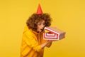 Portrait of surprised curious curly-haired woman with party cone hat looking into gift box, opening present