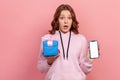 Portrait of surprised curly-haired teenage girl in hoodie holding smartphone and wrapped gift box, smiling to camera Royalty Free Stock Photo