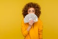 Portrait of surprised curly-haired woman in urban style hoodie peeking out of money with amazed eyes