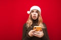Portrait of surprised girl in christmas hat and warm sweater stands on red background with smartphone in her hands and looks away Royalty Free Stock Photo