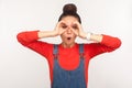 Portrait of surprised girl with hair bun in denim overalls looking through binoculars hand gesture, exploring discovering