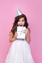 Portrait of surprised kid girl n birthday party princess hat holding gift box on a pink studio background Royalty Free Stock Photo