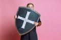 Portrait of surprised man, medieval warrior or archer in armor hiding himself behind a shield isolated over pink studio Royalty Free Stock Photo