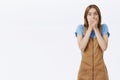 Portrait of surprised and shocked cute female model in brown dungarees gasping covering mouth with palms from amazement