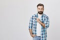 Portrait of suspicious bearded man pointing left with one hand and looking at camera, standing over gray background. Guy