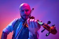 Portrait of talented young man, solo performer playing violin against gradient purple background in neon light Royalty Free Stock Photo