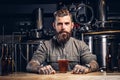 Portrait of a tattooed hipster male with stylish beard and hair in shirt sitting at the bar counter with glass of beer