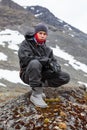 Portrait of Caucasian teenage boy dressing warm hiking outfit for mountaineering, young man sitting on big stone in mountains Royalty Free Stock Photo