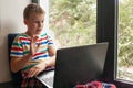 portrait of a teenage boy sitting on a windowsill with laptop on his lap, the end of home schooling, new normality. Royalty Free Stock Photo