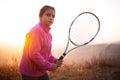 Portrait of teenage girl is training tennis skill outside court, in field at sunset. Wearing pink sweater and holding racket. Royalty Free Stock Photo