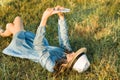 Portrait of teenage girl 14 years old lying on the grass. Girl in dress hat, in her headphones holds a smartphone, listens to Royalty Free Stock Photo