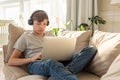 Portrait of teenager boy lying on a sofa in a room, wearing black headphones on his head, looks in laptop, on his knees. Royalty Free Stock Photo