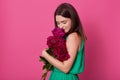 Portrait of tender beautiful young lady wearing in casual green dress, holding dark pink peonies, feeling smell of flowers, having