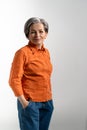 Portrait of tender mature woman wearing glasses looking at camera in orange shirt and denim pants. Pretty mid aged grey Royalty Free Stock Photo
