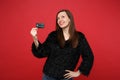 Portrait of thinkful young woman in black fur sweater looking up, holding credit bank card isolated on bright red