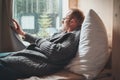 Portrait of thinking sad Middle-aged man in eyeglasses dressed open cardigan lying on cozy bed next to window looking out street