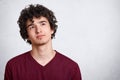 Portrait of thinking young man with dark curly hair, stands with thoughtful facial expression, dresses maroon shirt. Student