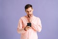 Portrait of thinking young man holding phone with doubtful and skeptical expression on pink  background. Royalty Free Stock Photo