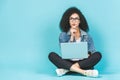 Portrait of thinking young afro american woman using laptop while sitting on a floor with legs crossed isolated over blue Royalty Free Stock Photo