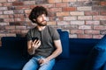 Portrait of thoughtful bearded young man looking away browsing wireless internet on mobile phone sitting on sofa at home Royalty Free Stock Photo