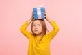 Portrait of thoughtful little girl holding gift on head and looking up with pensive face, curious about what`s inside box